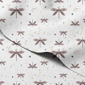 Könnte beinhalten: Weißer Stoff mit einem sich wiederholenden Muster aus braunen Libellen, kleinen schwarzen Sternen und goldenen Sprenkeln. Die Libellen sind gleichmäßig auf einem geometrischen Hintergrund verteilt. Der Stoff ist umgeschlagen und zeigt die Unterseite.