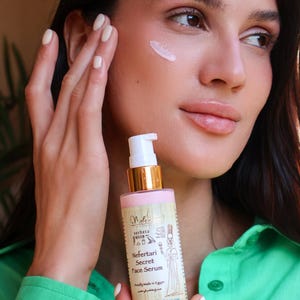 May include: A pink glass bottle with a gold cap and a white pump dispenser. The bottle is labeled "Nefertari Secret Face Serum" and features an image of an Egyptian queen. The bottle is being held by a person's hand.