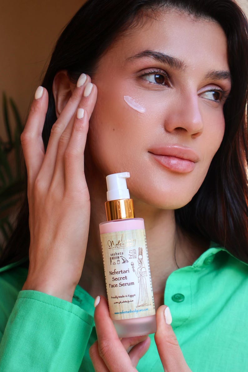 May include: A pink glass bottle with a gold cap and a white pump dispenser. The bottle is labeled "Nefertari Secret Face Serum" and features an image of an Egyptian queen. The bottle is being held by a person's hand.