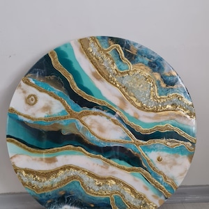 Puede incluir: Obra de arte abstracta redonda con un diseño en capas. La obra presenta tonos turquesa, blanco y azul marino, acentuados con purpurina dorada y resina. La pieza circular es un objeto decorativo.