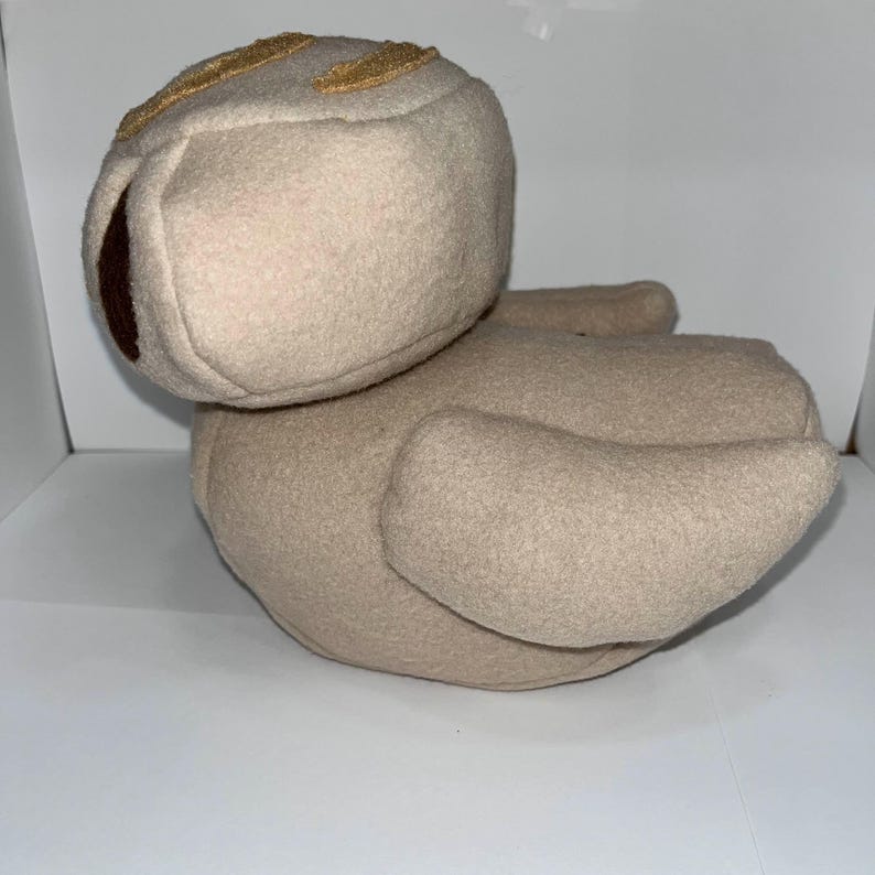 May include: A beige plush toy shaped like a duck. The duck has a large head with a brown eye and a gold-colored stitched design on top. The wings are outstretched, and the toy is sitting on a white surface.