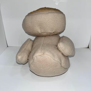 May include: A beige stuffed toy with a rounded head and small arms. The head has a gold-colored band around it. The toy is made of a soft, plush material and has a simple, handmade appearance.