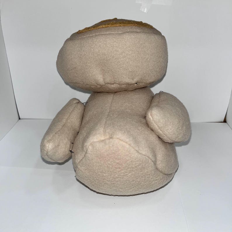 May include: A beige stuffed toy with a rounded head and small arms. The head has a gold-colored band around it. The toy is made of a soft, plush material and has a simple, handmade appearance.
