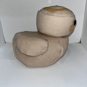 May include: A beige plush duck toy with a rounded body and head. The duck has a gold-colored accent on its head and small black eyes. The toy is made of soft fabric and is designed to resemble a duck.