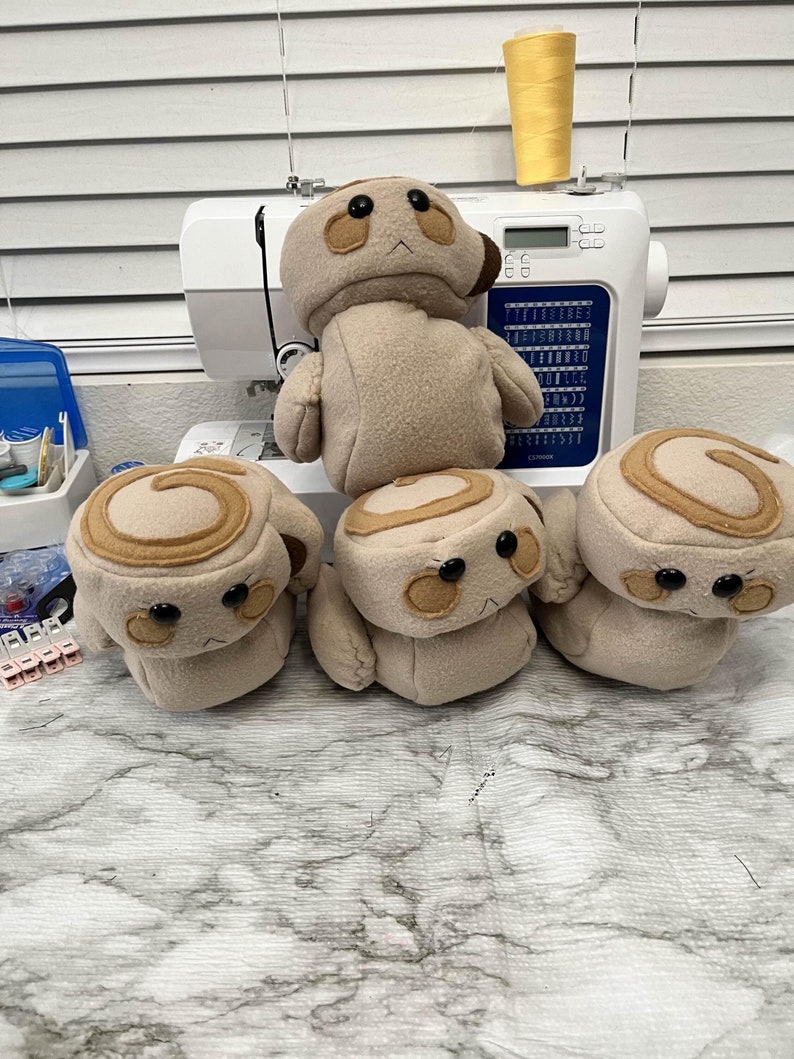 May include: A collection of handmade plush toys, including a larger figure and several smaller ones shaped like cinnamon rolls. The toys are beige with brown accents and black button eyes. A sewing machine and spool of yellow thread are in the background.