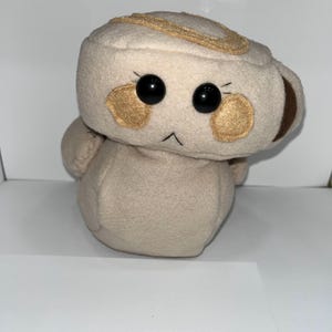 May include: A plush toy shaped like a coffee cup with a face. The cup is beige with brown accents and gold-colored details. It has large black eyes, blush-colored cheeks, and a stitched mouth.