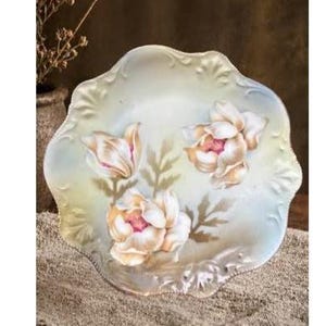 May include: A decorative ceramic plate with a scalloped edge and an iridescent finish. The surface features hand-painted floral motifs in shades of cream, soft pink, and muted green, depicting three blooming flowers with detailed petals and foliage.