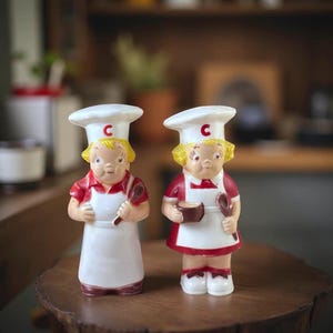 May include: Two vintage ceramic chef figurines. Each figurine wears a white chef's hat with a red "C", a red and white uniform, and a white apron. One holds a spoon, the other a bowl and spoon. The figurines are on a wooden surface.