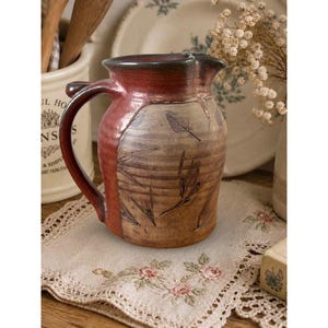 May include: A ceramic pitcher featuring a two-tone glaze with a deep reddish-brown top and a textured tan base. The body is decorated with dark, etched leaf patterns and horizontal ridges, complemented by a curved handle on the left side.
