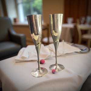 Puede incluir: Dos copas de champán plateadas con un diseño de tallo único, colocadas sobre un mantel blanco. Las copas tienen un acabado pulido y están cerca de dos pequeñas bayas rojas escarchadas. El fondo sugiere un restaurante.