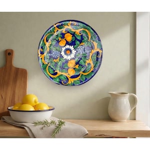 May include: A decorative ceramic plate with a vibrant design. The plate features a dark blue background with a floral and fruit pattern in green, yellow, and white. The plate is hanging on a light green wall, with a bowl of lemons and a pitcher in the foreground.