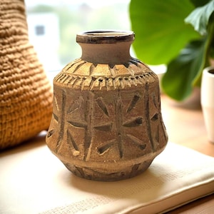 May include: A brown ceramic vase with a textured surface and geometric patterns. The vase has a wide base, a rounded body, and a narrow neck. The vase is approximately 7 inches tall.