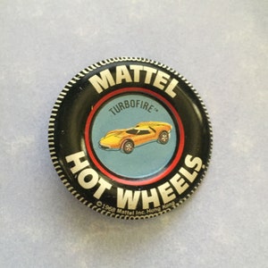 May include: A black and red Hot Wheels button with a yellow car and the text "MATTEL TURBOFIRE" and "1968 Mattel Inc. Hong Kong".