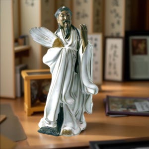 May include: A ceramic figurine of an Asian man in white robes, holding a fan. The figure has a long beard and is standing. The fan has gold accents. The figurine is on a wooden surface.