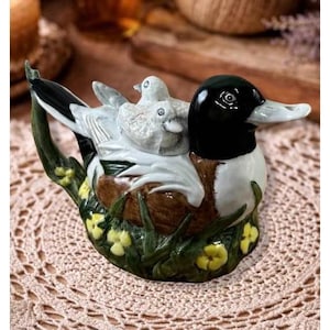 May include: A ceramic duck figurine with a black head, white body, and detailed wings. Two smaller ducklings sit on its back. The base features green grass, yellow flowers, and a brown nest. The figurine is on a lace doily.