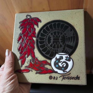 May include: Square ceramic tile with a Southwestern design. Features a cluster of red chili peppers, a black circular design, and a white pot with a lizard motif. The tile is tan with the text "83 Terssede".
