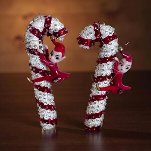 Puede incluir: Dos adornos de bastón de caramelo navideños. Cada bastón es blanco con rayas rojas y adornado con pequeños detalles brillantes. Un pequeño elfo con un atuendo rojo está unido a cada bastón de caramelo, como si lo estuviera escalando. El fondo es marrón oscuro.