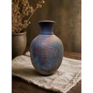 May include: A ceramic vase with a rounded body and a narrow neck, featuring a textured surface with a mottled finish in shades of deep blue, charcoal gray, and copper brown. The vessel has a smooth, dark rim and a weathered, rustic appearance.