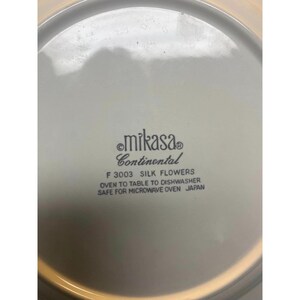 May include: Close-up of the bottom of a white ceramic plate with the text "mikasa Continental F 3003 SILK FLOWERS OVEN TO TABLE TO DISHWASHER SAFE FOR MICROWAVE OVEN JAPAN" printed in black.