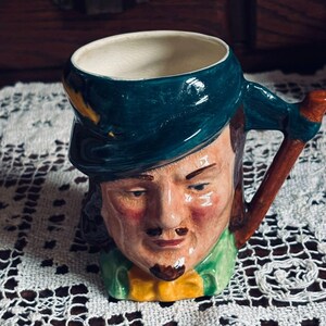 以下が含まれることがあります： 男性の頭の形をしたセラミックマグ。緑色の帽子と襟、茶色のハンドルが付いています。 マグには口ひげとあごひげのある顔が描かれており、黄色の蝶ネクタイが付いています。 白いレースのテーブルクロスの上に置かれています。