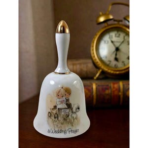 May include: A white porcelain bell with a gold-toned handle featuring a decorative illustration of two figures in a wagon with a dog. The text "a Wedding Prayer" is printed in black script at the bottom of the bell's exterior surface.