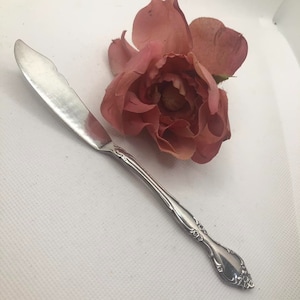 May include: A silver butter knife with a decorative handle and a rose with pink petals on a white surface.