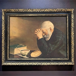 May include: Framed painting of an elderly person with a white beard, hands clasped in prayer. A loaf of bread, a bowl, and a book are on the table. The painting has a dark, ornate frame with gold accents.