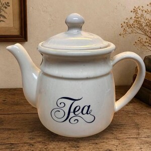 May include: A cream-colored ceramic teapot with a matching lid and a curved handle. The body of the teapot features the word Tea written in a dark blue, elegant cursive script. The surface has a subtle speckled texture throughout the ceramic finish.