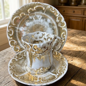 May include: A vintage teacup, saucer, and plate set. The set features a white and gold color scheme with ornate detailing. The teacup has a handle and sits on a matching saucer. The plate is in the background.