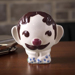 May include: A vintage ceramic figurine in the shape of a smiling clown's head. The clown has brown hair, a burgundy nose, and a dark brown mustache. The base of the figurine has blue polka dots. The figurine is on a wooden surface.
