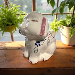 May include: A white ceramic dog ornament with closed eyes and pink accents on the ears and nose. The dog has blue and pink floral designs on its back. The ornament is on a wooden surface.