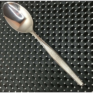 Op de afbeelding: Een zilveren lepel met een getextureerd handvat. De lepel ligt op een zwart-wit geweven placemat.