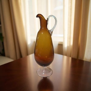 Könnte beinhalten: Eine Vintage-Karaffe aus bernsteinfarbenem Glas mit einem klaren Griff und Sockel. Die Karaffe hat einen langen, schlanken Hals und einen abgerundeten Körper. Sie steht auf einem dunklen Holztisch, mit einem Fenster und Vorhängen im Hintergrund.