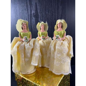 May include: Three vintage figurines of women in yellow dresses and hats, holding bouquets. The dresses have a lace overlay, and the bodices are green. The figurines are on a gold-colored surface.