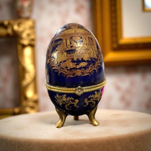 May include: A decorative, cobalt blue egg-shaped trinket box with gold accents. The egg features a detailed gold illustration of figures in a scene. The box has a gold band around the middle and rests on three gold feet. A unique collectible.