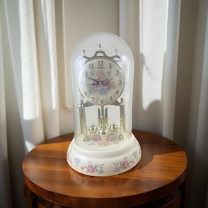 May include: A Timex anniversary clock under a clear glass dome. The clock face features black numerals and a floral pattern in pink and blue. The base is cream-colored with a matching floral design, and the internal mechanism is gold-toned metal.