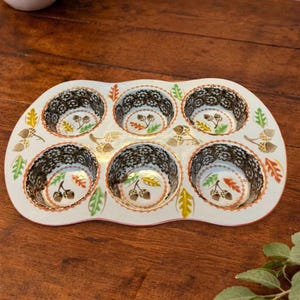 May include: A white ceramic muffin pan with six circular cavities, featuring a decorative pattern of brown acorns and multi-colored autumn leaves in shades of orange, yellow, and green, with a dark brown lace-like border around each cup.