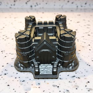 May include: A dark gray castle-shaped cake pan with four turrets and a central gatehouse. The pan has detailed architectural features, including arched doorways and crenellations, and is made of a non-stick material.