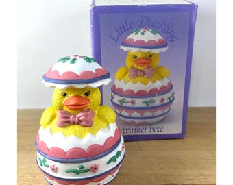 May include: A yellow duckling peeking out of a pink and white egg-shaped trinket box. The box has a lid that opens to reveal the duckling inside. The box is decorated with pink and white stripes, flowers, and green leaves. The box is labeled "Little Duckling Trinket Box".