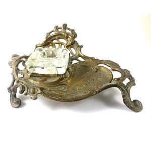 May include: An antique brass ashtray with a clear glass square ashtray insert. The brass base has an ornate, floral design. The ashtray is on a white background.