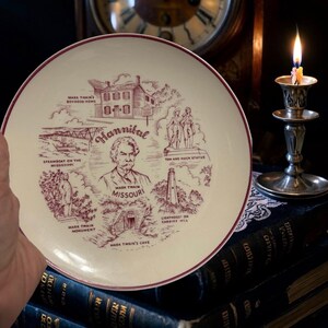 Op de afbeelding: Een crèmekleurige souvenirbord met een bordeauxrode rand en illustraties van bezienswaardigheden in Hannibal, Missouri, waaronder het jeugdhuis van Mark Twain, een raderstoomboot en een vuurtoren. Het bord bevat de tekst "Hannibal" en "Mark Twain Missouri".