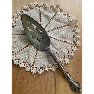 Può includere: Un servizio per torta in argento con un design elaborato e un manico decorativo. Il servizio è appoggiato su un tovagliolo di pizzo bianco all'uncinetto.