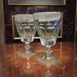 Puede incluir: Dos copas de vino de cristal tallado transparente. Cada copa tiene una base redonda, un tallo con un bulbo decorativo y una copa con un patrón geométrico. Las copas están sobre una superficie marrón rojiza, con un espejo enmarcado al fondo.