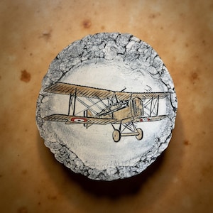 May include: A hand-painted, round ceramic plate featuring a vintage biplane design. The biplane is depicted in tan and brown with red and white accents, set against a white background. The plate's edge has a textured, gray and white border.