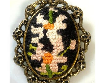 May include: Vintage oval brooch with a floral design. The brooch features a black background with a detailed embroidered flower in white, pink, orange, and green. The brooch is framed by a gold-toned, ornate metal border.