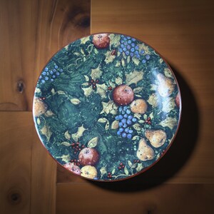 May include: Circular decorative plate with a green backdrop, adorned with apples, pears, grapes, and holly leaves. The plate has a red rim and is placed on a wooden surface.