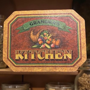 Puede incluir: Un letrero rectangular de madera con un borde decorativo. El letrero presenta una pancarta que dice "Grandma's" encima de una cornucopia llena de frutas y verduras. La palabra "KITCHEN" se muestra en letras doradas grandes.