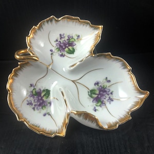 May include: A white porcelain leaf-shaped dish with three sections. The dish is decorated with purple and green flowers and has a gold rim.
