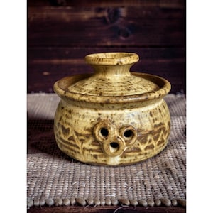 May include: A handcrafted, ceramic butter dish with a lid. The dish is a mottled tan color with a textured surface and decorative cutouts. The lid has a small knob for easy removal. The dish sits on a woven mat.