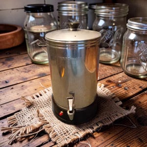Puede incluir: Una cafetera cromada vintage con base y grifo negros, sobre un posavasos de arpillera. La cafetera tiene tapa y asas, con la marca "MIRRO-MATIC" visible. Varias jarras de vidrio están en el fondo, todo sobre una superficie de madera.